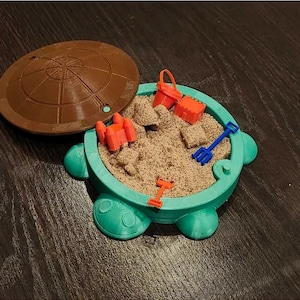 May include: A teal plastic sandbox with a brown lid. The sandbox is filled with sand and has a small orange castle, a blue shovel, and a red bucket inside.