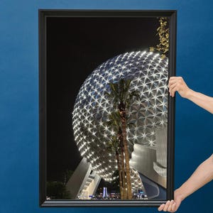 Peut inclure: Une photographie encadrée en noir d'un grand dôme géodésique illuminé la nuit. Le dôme est composé de nombreux petits panneaux triangulaires et est éclairé de lumières blanches. Deux palmiers se trouvent au premier plan.