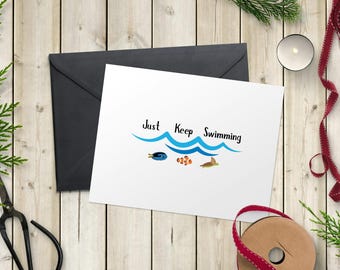 Puede incluir: Una tarjeta blanca con el texto "Just Keep Swimming" y una ilustración de olas y peces, junto a un sobre negro. La tarjeta está sobre una superficie de madera con elementos decorativos.