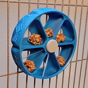May include: A blue pet treat dispenser in the shape of a wheel, with six compartments containing small, round treats. The dispenser is mounted on a metal cage. The wheel has a textured design on the outer edge and a white centre.