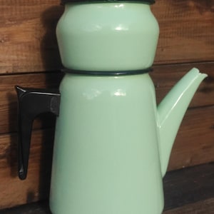 Op de afbeelding: Een mintgroene emaille koffiepot met een zwarte handgreep, deksel en afwerking. De pot in vintage-stijl heeft een klassiek ontwerp met een tuit en een deksel met een zwarte knop. De pot wordt waarschijnlijk gebruikt voor het zetten van koffie of thee.