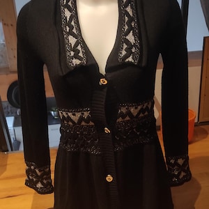Puede incluir: Cárdigan negro con adornos decorativos plateados y dorados en el cuello, la cintura y los puños. El suéter tiene cuello en V, mangas largas y botones dorados. La parte inferior del cárdigan se ensancha ligeramente.