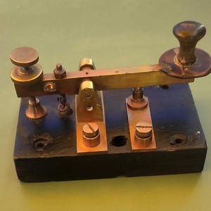 May include: A brass telegraph key mounted on a rectangular black wooden base. The device features a horizontal metal arm with a circular knob at one end, an adjustable screw mechanism at the other, and a central pivot support secured by two screws.