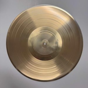 Puede incluir: Un disco de vinilo de color dorado con un agujero central y surcos concéntricos. El disco está limpio y sin rayones.