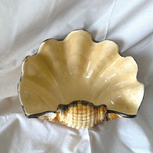 May include: A ceramic seashell-shaped dish with a cream-colored interior and a black-lined edge. The base of the shell is textured with brown and tan hues. The dish is designed to resemble a scallop shell.