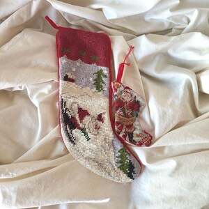 May include: A large needlepoint Christmas stocking featuring a winter landscape with a red top border and a smaller matching needlepoint ornament shaped like a stocking, both with red hanging loops and detailed stitched imagery.