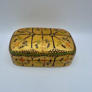 May include: A rectangular box with rounded corners featuring a gold-toned base color. The surface is decorated with black line work forming a grid pattern, accented by painted floral motifs in shades of orange, green, and black throughout.