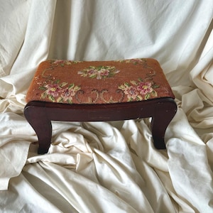 May include: A rectangular footstool featuring a dark brown wooden frame with four curved legs. The top surface is upholstered in a textured needlepoint fabric with a burnt orange background and a floral pattern in shades of pink, green, and white.