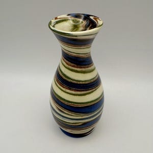 May include: A decorative ceramic vase with a striped pattern. The vase features alternating bands of blue, white, green, and brown. The vase has a rounded body and a flared opening. The vase is approximately 7 inches tall.