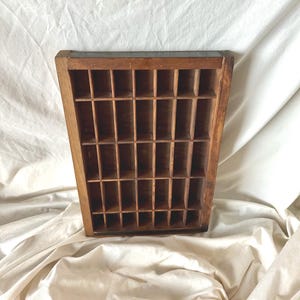 May include: A rectangular wooden printer's tray featuring a grid of multiple small, open compartments. The tray is constructed from dark brown wood with visible grain and has a sturdy frame surrounding the interior dividers.