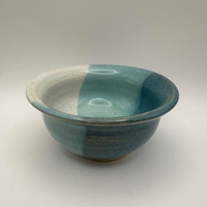 May include: A handmade ceramic bowl with a two-tone design. The bowl features a white and teal color scheme, with a darker teal on the exterior. The bowl has a rounded shape and a wide rim.