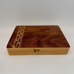 May include: A rectangular wooden box with a dark brown stained top and a lighter wood base. The top features an etched decorative border. A small metal clasp secures the box.