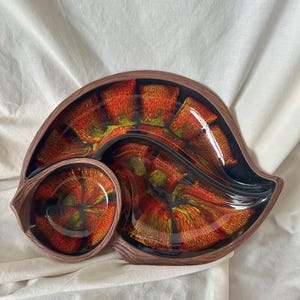 May include: A ceramic serving dish with a shell-like shape, featuring a glossy finish with a vibrant red, orange, and yellow sunburst pattern against a black background. The dish has a brown, textured rim that mimics the appearance of wood grain.