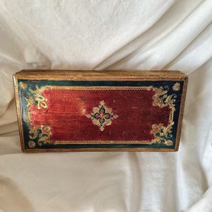 May include: Rectangular wooden box with a red surface and gold trim. The box features ornate gold and green corner designs and a central floral pattern. The box is likely used for storage or display.