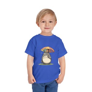 Op de afbeelding: Blauw peuter T-shirt met een cartoon illustratie van een grijs personage dat een regenboog paraplu vasthoudt. Het personage heeft een witte buik en staat op groen gras. Een gele ster is zichtbaar.