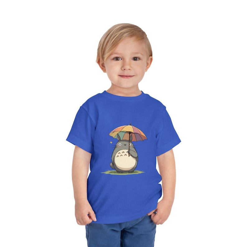 Op de afbeelding: Blauw peuter T-shirt met een cartoon illustratie van een grijs personage dat een regenboog paraplu vasthoudt. Het personage heeft een witte buik en staat op groen gras. Een gele ster is zichtbaar.