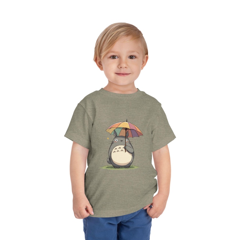 Op de afbeelding: Een peuter draagt een heidegroen t-shirt met een cartoon Totoro-personage met een kleurrijke paraplu. De paraplu heeft regenboogkleurige panelen. Het Totoro-personage is grijs met een witte buik en staat op groen gras.