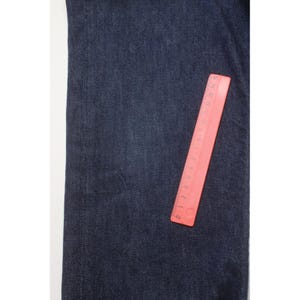 May include: A pair of dark blue denim jeans featuring a visible weave texture. A red plastic ruler is placed vertically on the fabric, showing measurements in inches and centimeters, with the ruler length spanning approximately 6 inches.