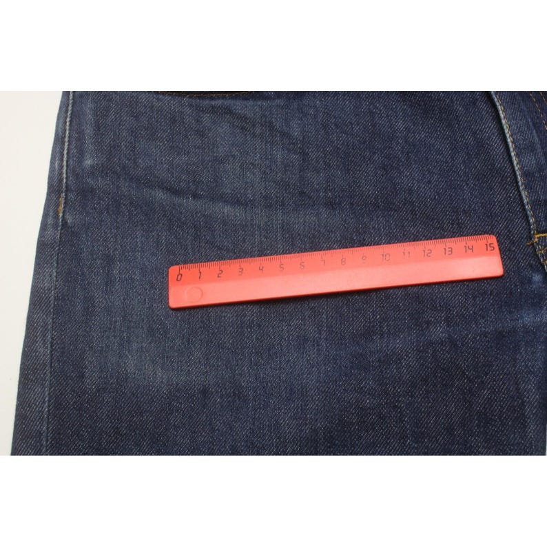 May include: Dark blue denim fabric with visible stitching and a textured weave. A red plastic ruler is placed horizontally across the fabric, showing a measurement scale from 0 to 15 centimeters, which is approximately 5.9 inches in length.