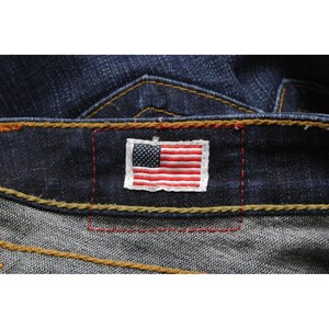 May include: Dark blue denim jeans featuring a rectangular patch with an embroidered American flag on the waistband. The jeans are constructed with thick, contrasting gold-colored stitching and red thread accents around the flag patch.