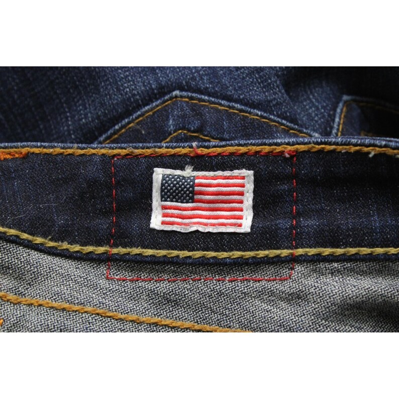 May include: Dark blue denim jeans featuring a rectangular patch with an embroidered American flag on the waistband. The jeans are constructed with thick, contrasting gold-colored stitching and red thread accents around the flag patch.