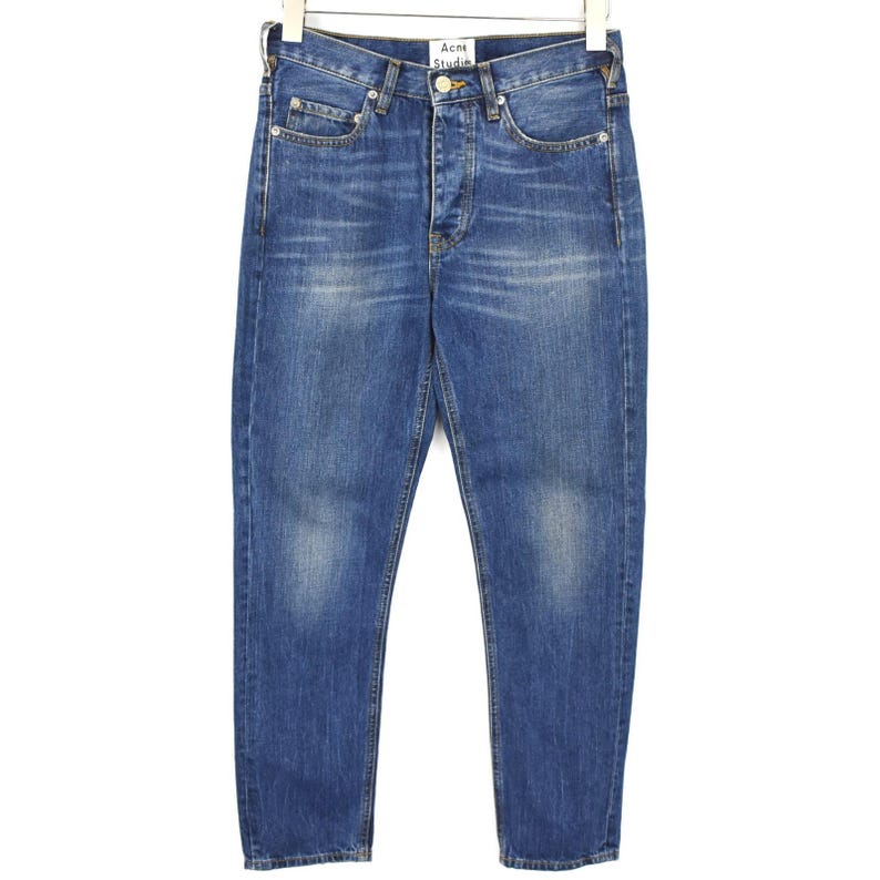 May include: A pair of blue denim jeans with a faded wash, featuring a button fly closure, five pockets, and a white brand label reading Acne Studios at the interior waistband. The jeans have a straight leg cut with visible stitching details.