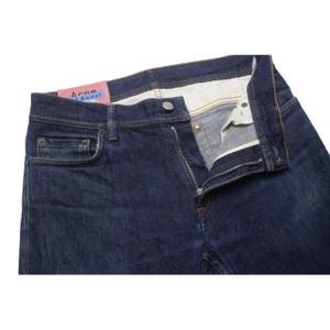 May include: Dark blue denim jeans featuring a metal button closure, a zippered fly, and five pockets. A pink rectangular label with the text Acne Studios Bl&aring; Konst is attached to the interior waistband. The interior pocket lining is off-white.