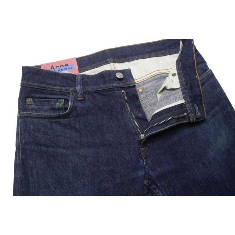 May include: Dark blue denim jeans featuring a metal button closure, a zippered fly, and five pockets. A pink rectangular label with the text Acne Studios Bl&aring; Konst is attached to the interior waistband. The interior pocket lining is off-white.