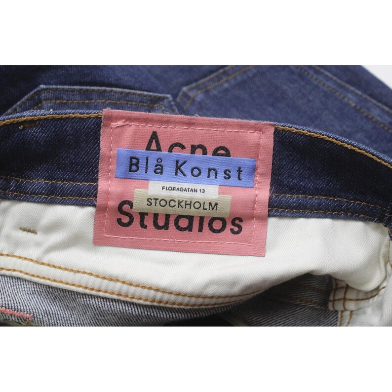 May include: Dark blue denim jeans featuring a rectangular pink brand label stitched onto the interior waistband. The label displays the text Acne Bl&aring; Konst, Floragatan 13, and Stockholm Studios in black and blue lettering on a pink background.