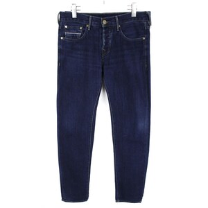 May include: Dark blue denim jeans featuring a classic five-pocket design, a button fly closure, and metallic rivets. The pants have a straight-leg cut with subtle fading on the thighs and are suspended from a metal hanger for display purposes.