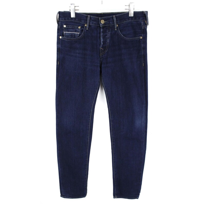 May include: Dark blue denim jeans featuring a classic five-pocket design, a button fly closure, and metallic rivets. The pants have a straight-leg cut with subtle fading on the thighs and are suspended from a metal hanger for display purposes.