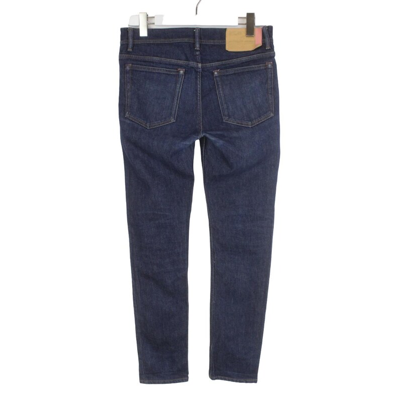 May include: Dark blue denim jeans featuring a classic five-pocket design with metal rivets at the corners of the back pockets. A rectangular tan leather-like patch is stitched onto the back waistband, and the garment is suspended from a hanger.