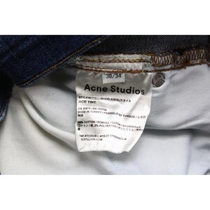 May include: A pair of dark blue denim jeans featuring an interior white fabric care label. The label displays the text Acne Studios, Ace Tint, 30/34, Made in Turkey, and indicates a composition of 98 percent cotton and 2 percent polyester.