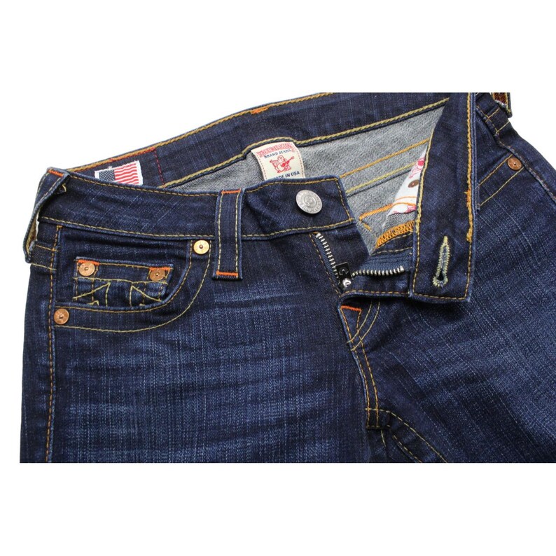May include: Dark blue denim jeans featuring thick orange contrast stitching, a metal button fly, and a small coin pocket. The interior waistband includes a True Religion Brand Jeans label and a small American flag tag on the inner lining.