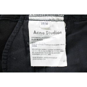 May include: A black garment interior featuring a white rectangular care label with the text Acne Studios, FLEX BLACK, 27/32, and material composition of 92% cotton, 5% polyester, and 3% elastane, with manufacturing details indicating made in Albania.