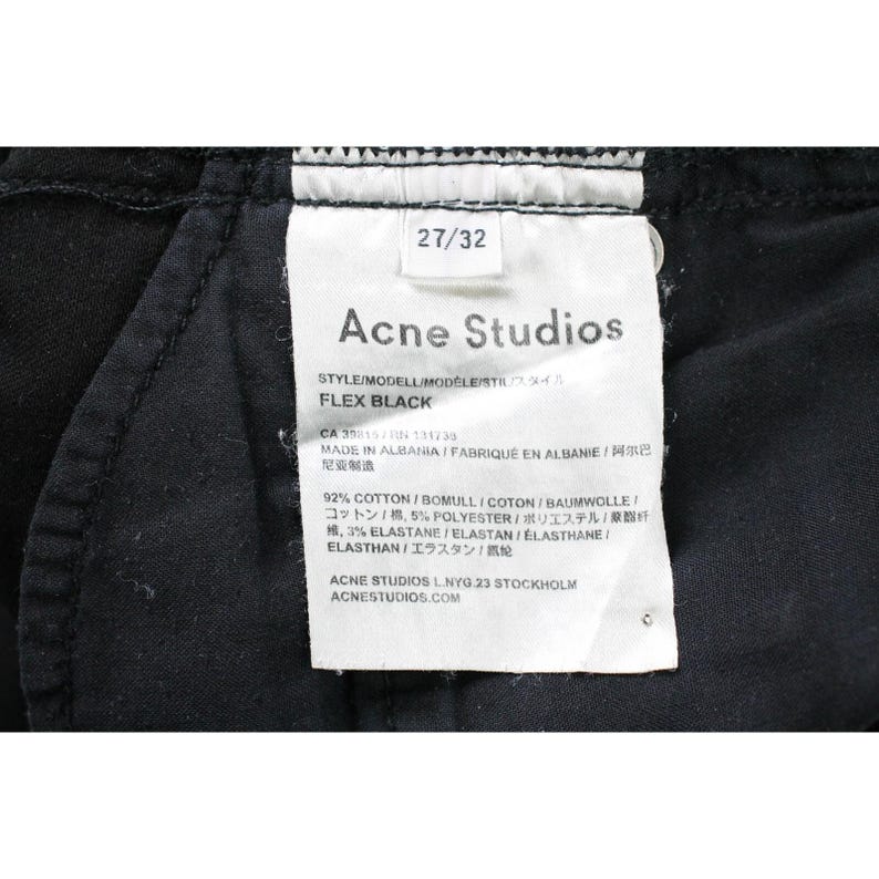 May include: A black garment interior featuring a white rectangular care label with the text Acne Studios, FLEX BLACK, 27/32, and material composition of 92% cotton, 5% polyester, and 3% elastane, with manufacturing details indicating made in Albania.