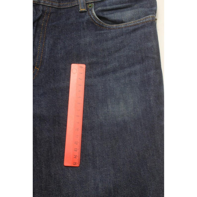May include: Dark blue denim jeans featuring a classic five-pocket construction with visible stitching and a metal button closure. A red plastic ruler is placed vertically on the fabric, showing a measurement scale of approximately 6 inches in length.