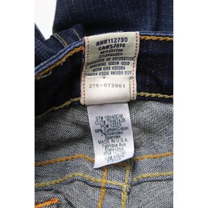 May include: Dark blue denim jeans featuring yellow contrast stitching and two white fabric care labels. The labels display text including RN#112790, CA#57414, 98% cotton, 2% spandex, ST# 10H40OM, CUT# TRW7428, PO# 12373, MADE IN U.S.A., and the style name STELLA.