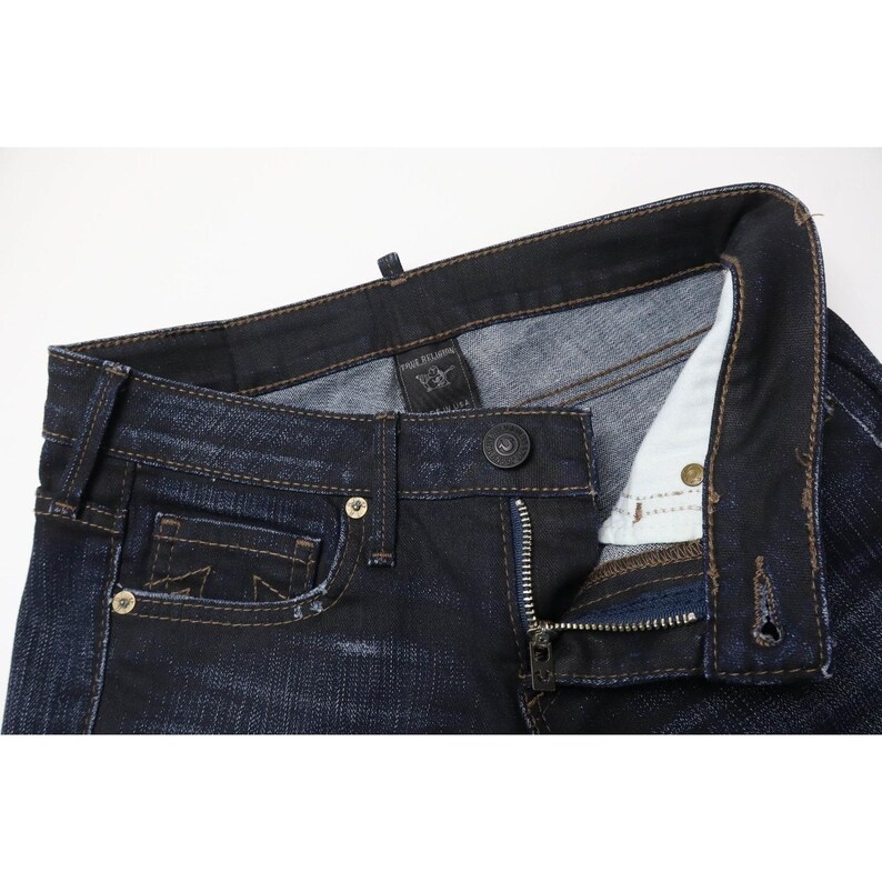 Peut inclure: Jean en denim bleu indigo fonc&eacute; avec une fermeture &agrave; boutons, des rivets en m&eacute;tal cuivr&eacute; et une petite poche gousset. L'int&eacute;rieur de la ceinture pr&eacute;sente une &eacute;tiquette noire de la marque True Religion. Le tissu pr&eacute;sente un d&eacute;lavage subtil.