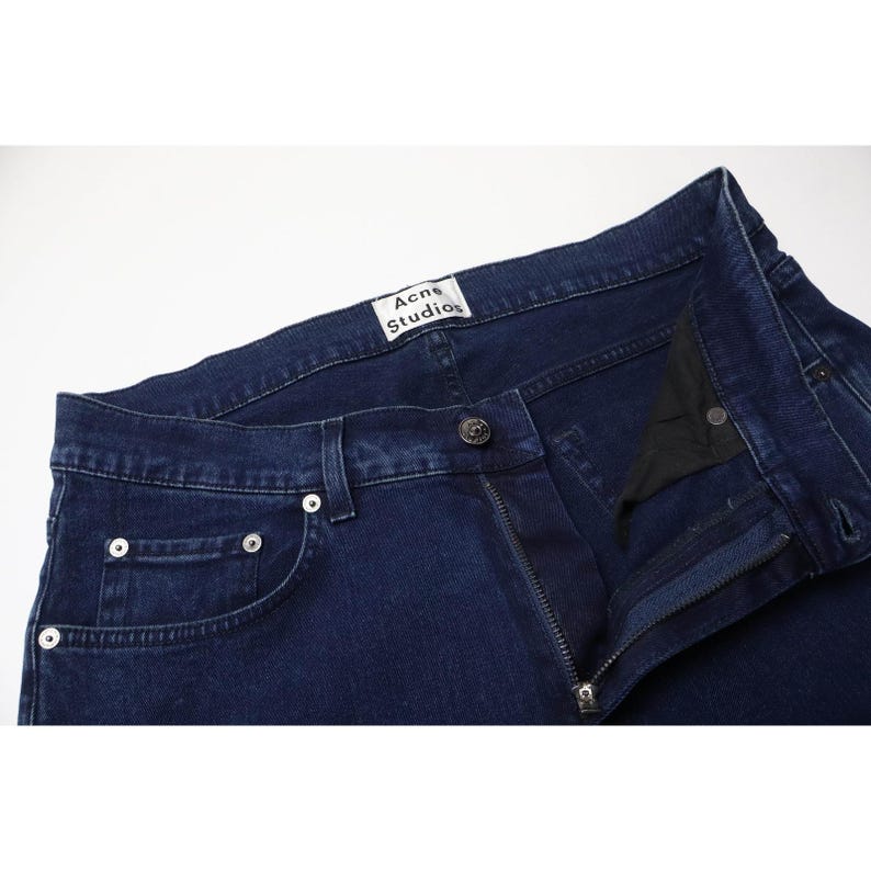 Pu&ograve; includere: Jeans in denim blu scuro con chiusura a bottoni in metallo, rivetti color argento e un'etichetta bianca all'interno della cintura con il testo nero Acne Studios. I jeans includono un taschino portamonete e passanti per cintura lungo il bordo.