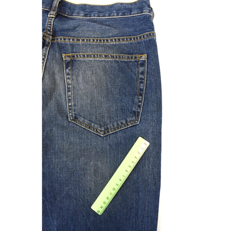 May include: A pair of blue denim jeans featuring a classic five-pocket design with a single visible back pocket and yellow contrast stitching. A green plastic ruler measuring 15 centimetres is placed diagonally across the fabric for scale.