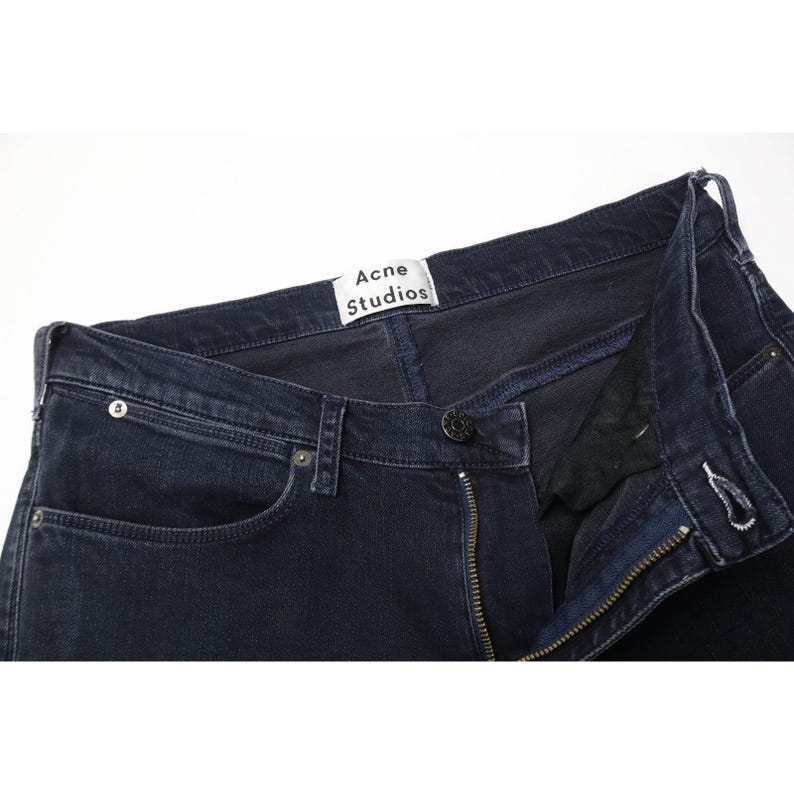 Pode incluir: Cal&ccedil;as de ganga em azul &iacute;ndigo escuro com design cl&aacute;ssico de cinco bolsos, fecho de bot&otilde;es met&aacute;licos e uma etiqueta branca no interior do c&oacute;s com o texto preto Acne Studios. As cal&ccedil;as apresentam costuras tonais e rebites de metal prateado.