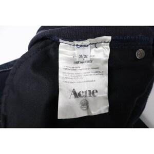 Pode incluir: Um par de cal&ccedil;as de ganga azul-escuras com uma etiqueta de cuidados interior em tecido branco. A etiqueta apresenta o texto Acne, Max Man Ray, 31/32, Made in Turkey, 98% algod&atilde;o e 2% elastano. Um rebite de metal prateado &eacute; vis&iacute;vel.