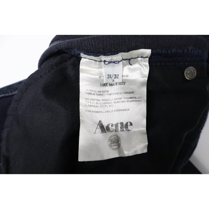 Pode incluir: Um par de cal&ccedil;as de ganga azul-escuras com uma etiqueta de cuidados interior em tecido branco. A etiqueta apresenta o texto Acne, Max Man Ray, 31/32, Made in Turkey, 98% algod&atilde;o e 2% elastano. Um rebite de metal prateado &eacute; vis&iacute;vel.