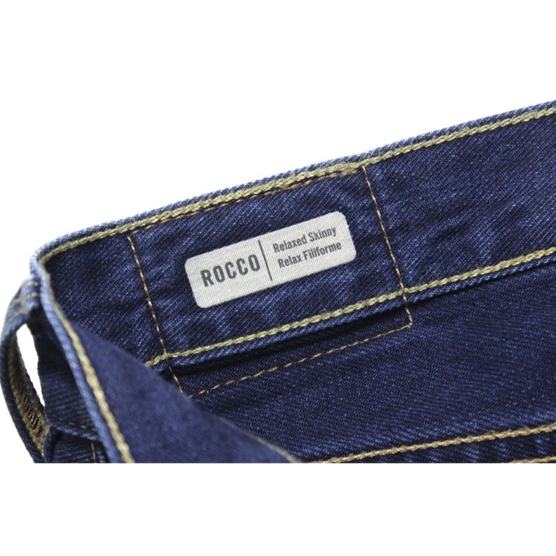 May include: Dark blue denim jeans featuring a white interior label with the text ROCCO Relaxed Skinny Relax Filiforme. The fabric has a visible twill texture and is constructed with thick, contrasting gold-colored stitching along the waistband.