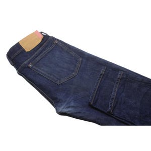 May include: Dark indigo blue denim jeans featuring a rectangular tan leather patch on the back waistband with a small pink accent tab. The jeans have a classic five-pocket construction with visible stitching, metal rivets, and a folded leg hem.
