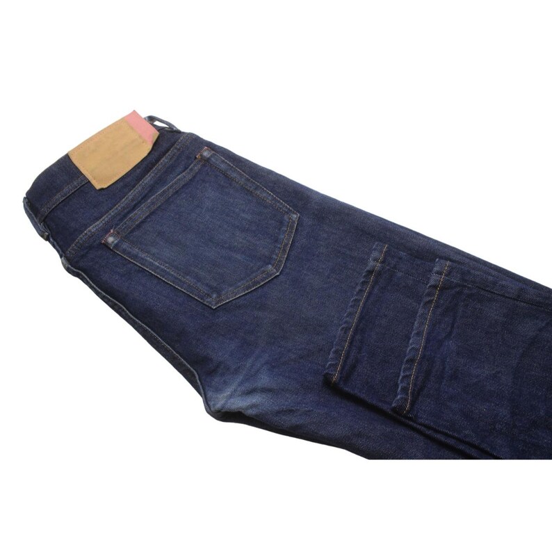 May include: Dark indigo blue denim jeans featuring a rectangular tan leather patch on the back waistband with a small pink accent tab. The jeans have a classic five-pocket construction with visible stitching, metal rivets, and a folded leg hem.