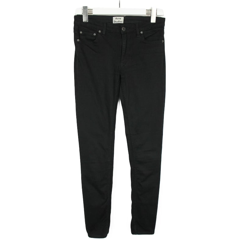 May include: A pair of solid black denim jeans featuring a classic five-pocket design, a button and zipper fly closure, and a white brand label reading Acne Studios stitched onto the interior waistband, all displayed on a white clothing hanger.
