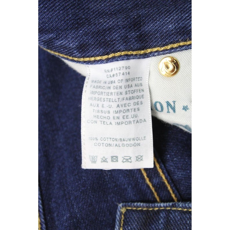 May include: A dark blue denim garment featuring a white interior care label with black text detailing manufacturing information, 100% cotton composition, and laundry symbols. A gold-toned metal rivet with a horseshoe logo is visible nearby.