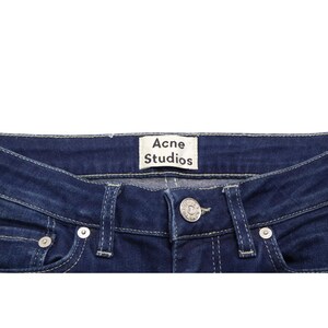 Puede incluir: Vaqueros de mezclilla azul oscuro con una etiqueta de marca rectangular blanca con el texto Acne Studios cosida en la cintura interior. Los vaqueros incluyen un cierre de bot&oacute;n de metal plateado, trabillas para cintur&oacute;n y costuras en contraste.